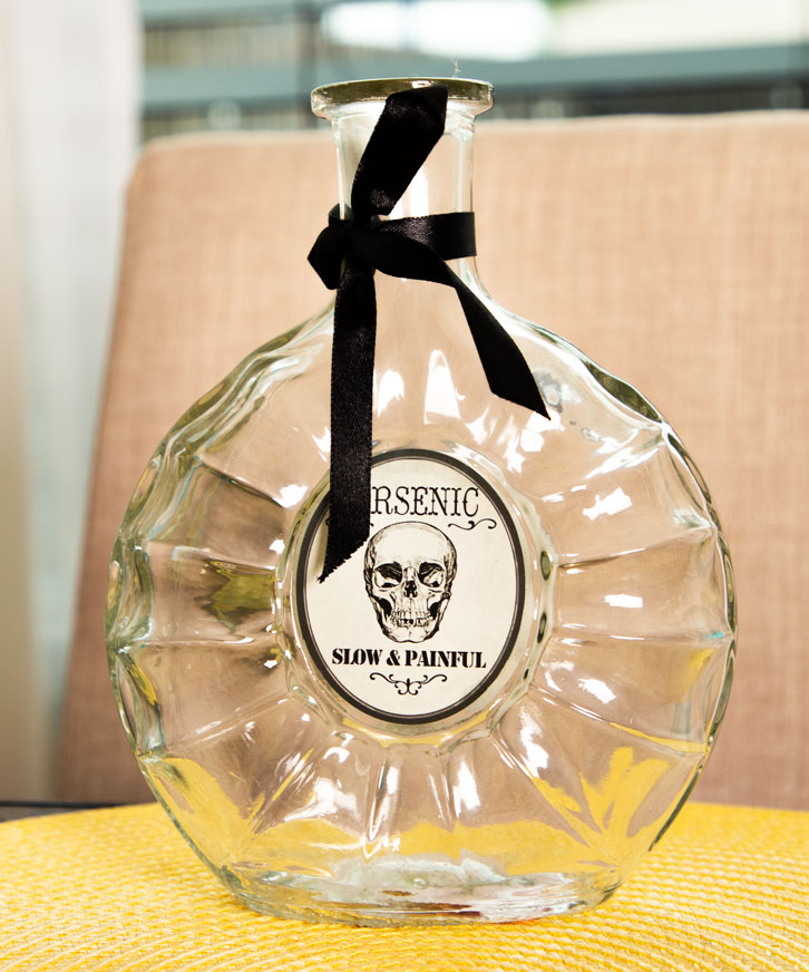 A clear glass arsenic bottle with a black skull-shaped cap and a label that reads 'Resinic - Slow & Painful'.