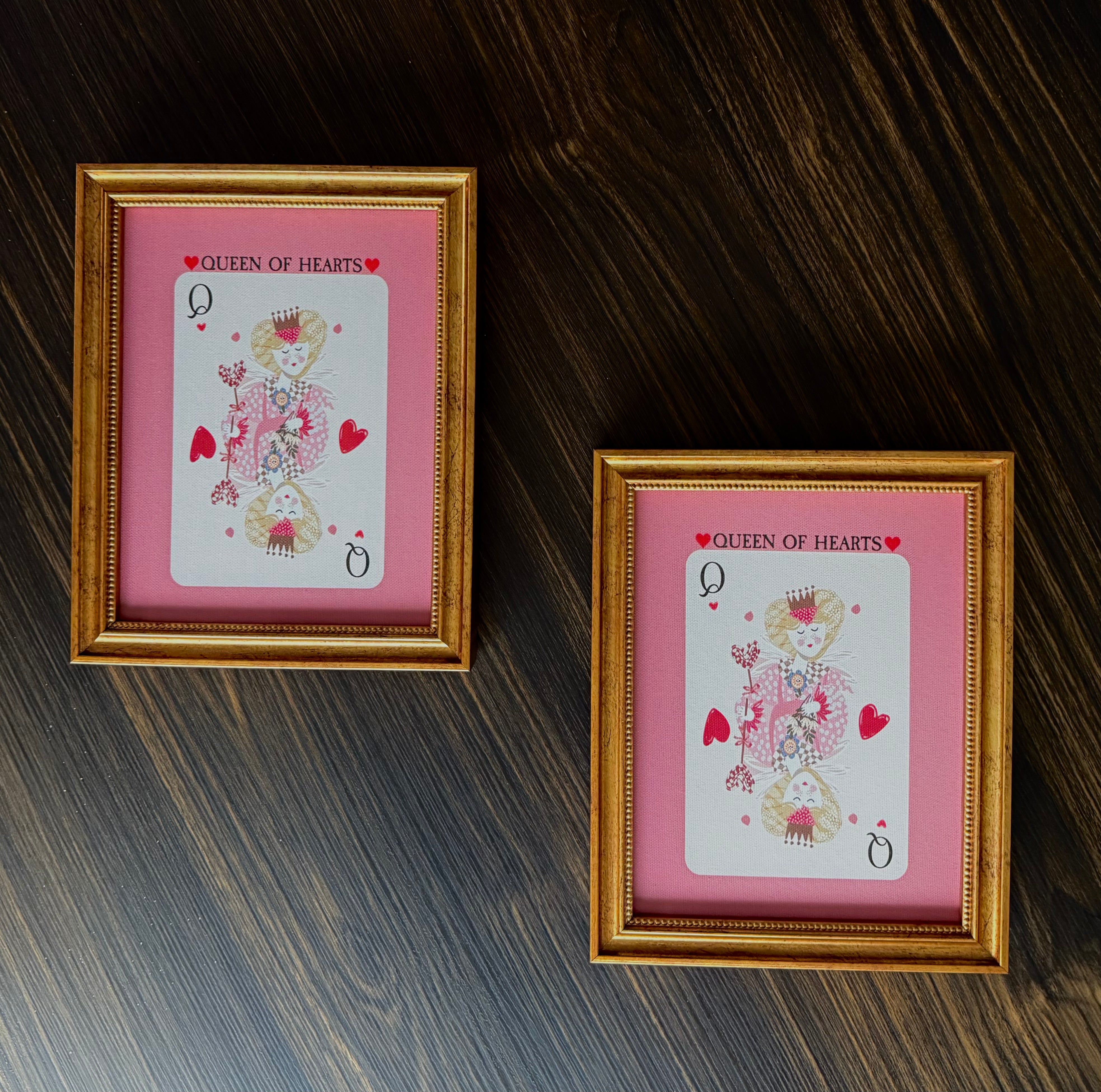 Two framed illustrations of the 'Queen of Hearts' from a deck of cards on a wooden surface.