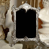 Decorative silver frame with black center on a reflective surface with classical sculptures in the background.