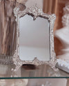 Decorative silver mirror with intricate frame on a glass surface