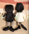 Two black hooded dolls with striped socks and green gloves on a textured surface.
