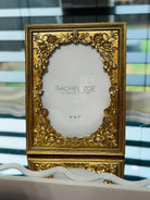 Ornate baroque gold rectangular picture frame with raised floral corners, 5x7 opening, displayed on a mirrored vanity.