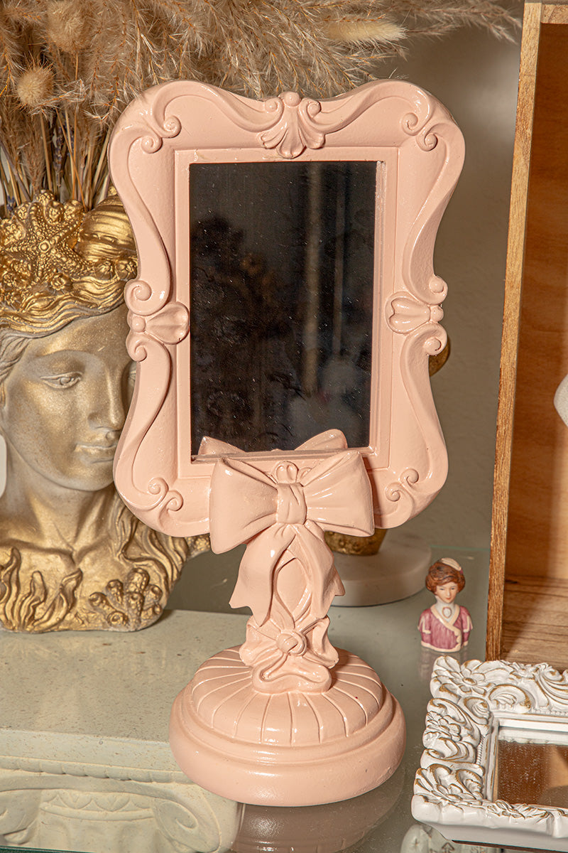 Blush pink vanity mirror with ornate bow design, shabby chic accent for vintage vanity or bedroom decor.
