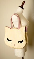 A beige straw toe handbag with a cat design, featuring button closure and a mannequin display. Hello Kitty inspired bag.